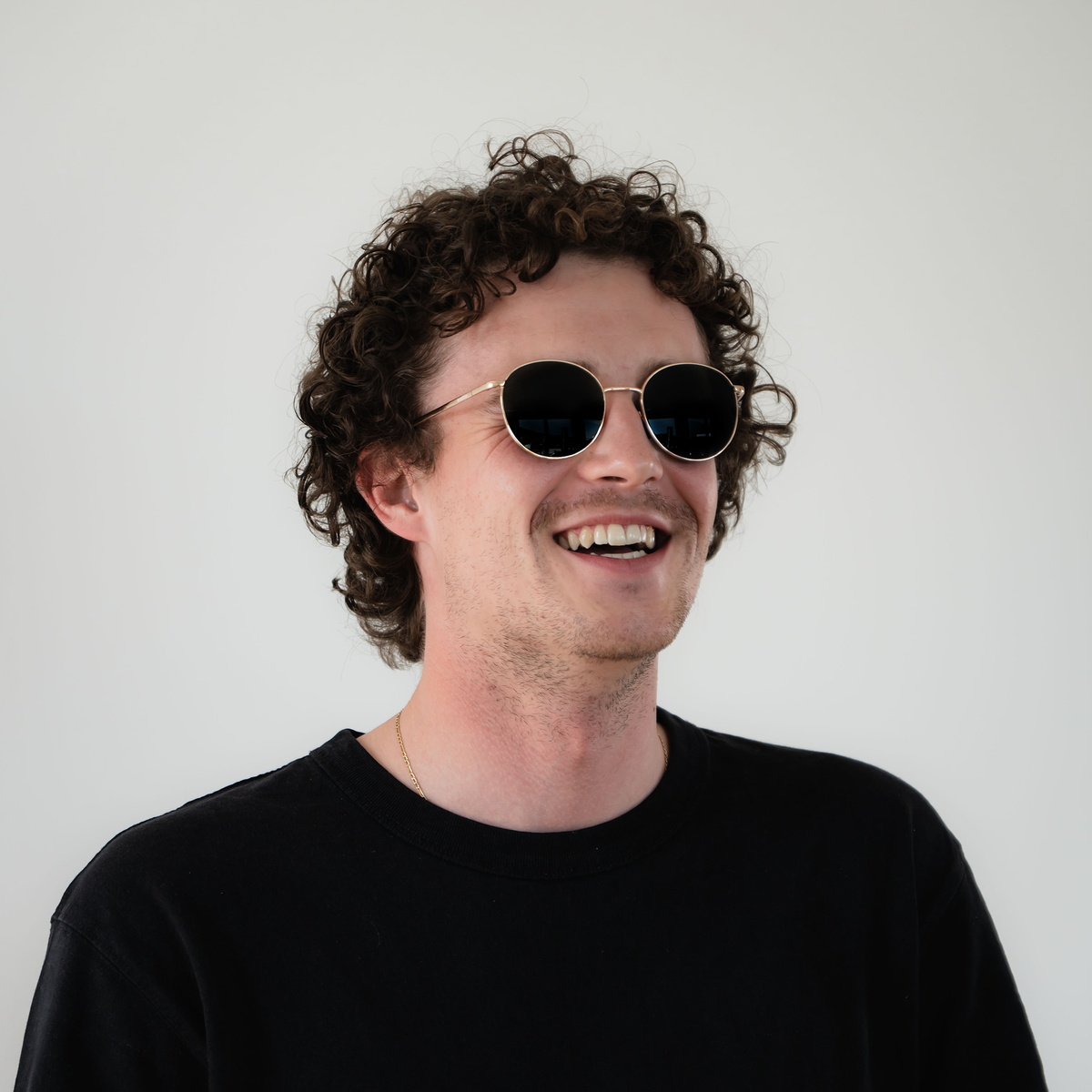 A man with curly hair and sunglasses smiles, wearing a black shirt against a plain white background. He sports the stylish NAKED Optics The PARKER Metallrahmen Sonnenbrille, which offers reliable UV400 Schutz.