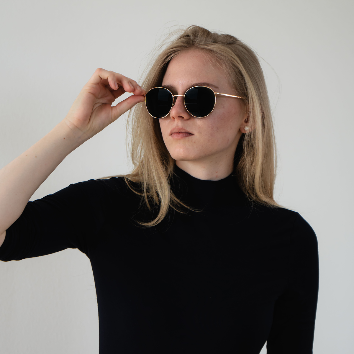 A woman in a black turtleneck and sunglasses touches her glasses while posing against a plain white background. She is wearing “The PARKER” by NAKED Optics, featuring a metal frame and UV400 protection for timeless style and optimal eye safety.