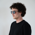 A man with curly hair and sunglasses wearing a black shirt stands against a plain white background, effortlessly showcasing the stylish NAKED Optics The PARKER sunglasses featuring UV400 protection.