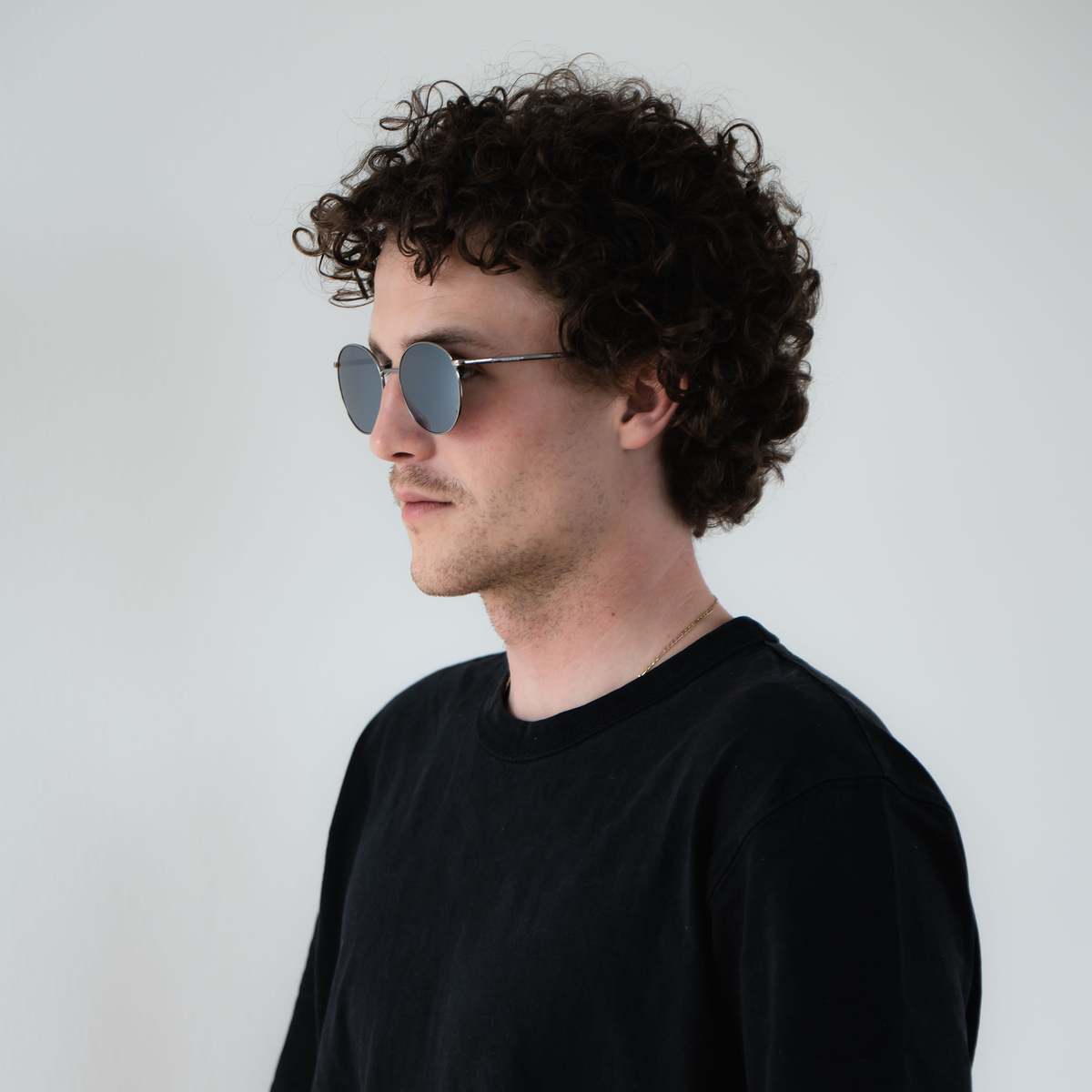 A man with curly hair and sunglasses wearing a black shirt stands against a plain white background, effortlessly showcasing the stylish NAKED Optics The PARKER sunglasses featuring UV400 protection.