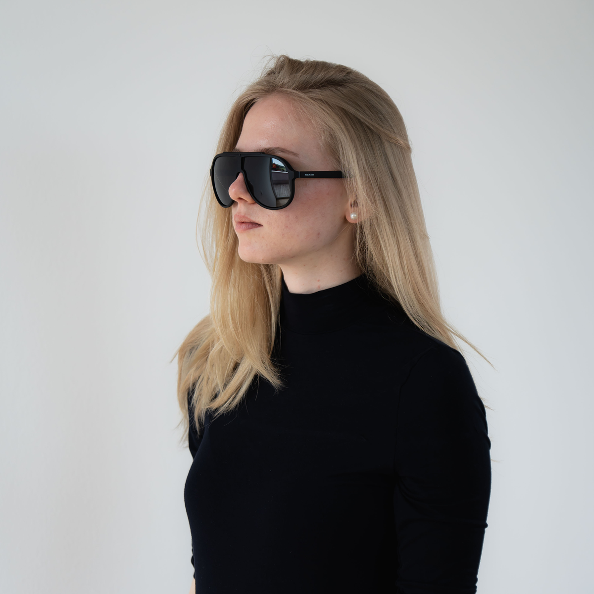 Woman with long blonde hair wearing NAKED Optics The PILOT UV400 black sunglasses and a black turtleneck against a plain background.