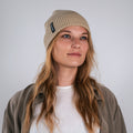Una donna con un berretto beige NAKED Optics Beanie POLAR e abiti casual guarda leggermente verso l'alto, in piedi davanti a uno sfondo chiaro e uniforme.