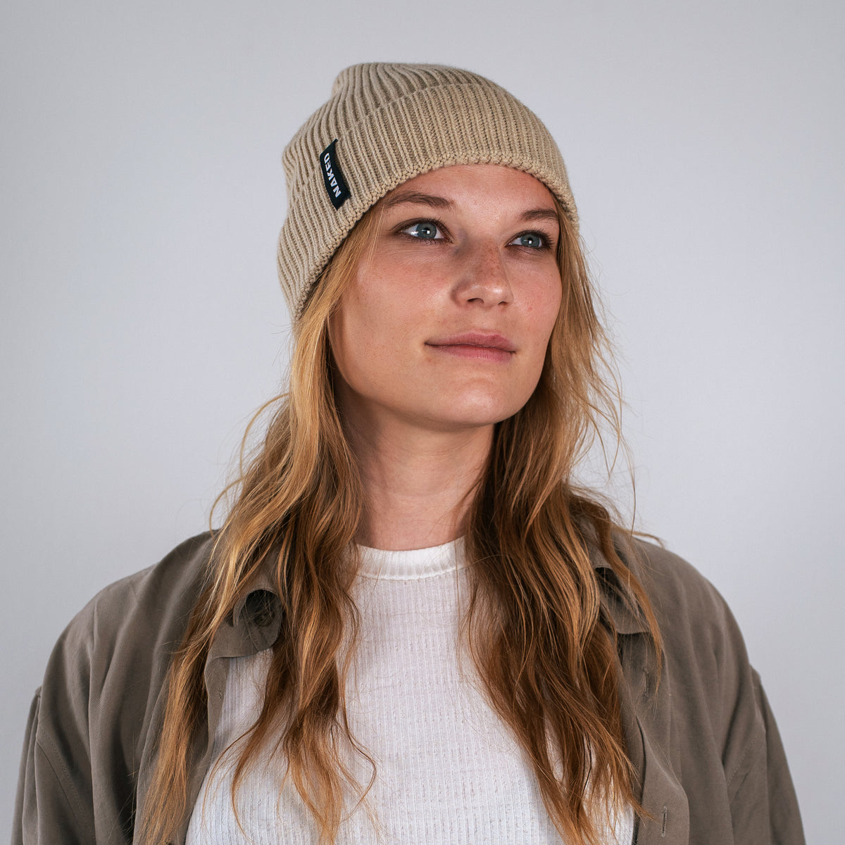 Una donna con un berretto beige NAKED Optics Beanie POLAR e abiti casual guarda leggermente verso l'alto, in piedi davanti a uno sfondo chiaro e uniforme.