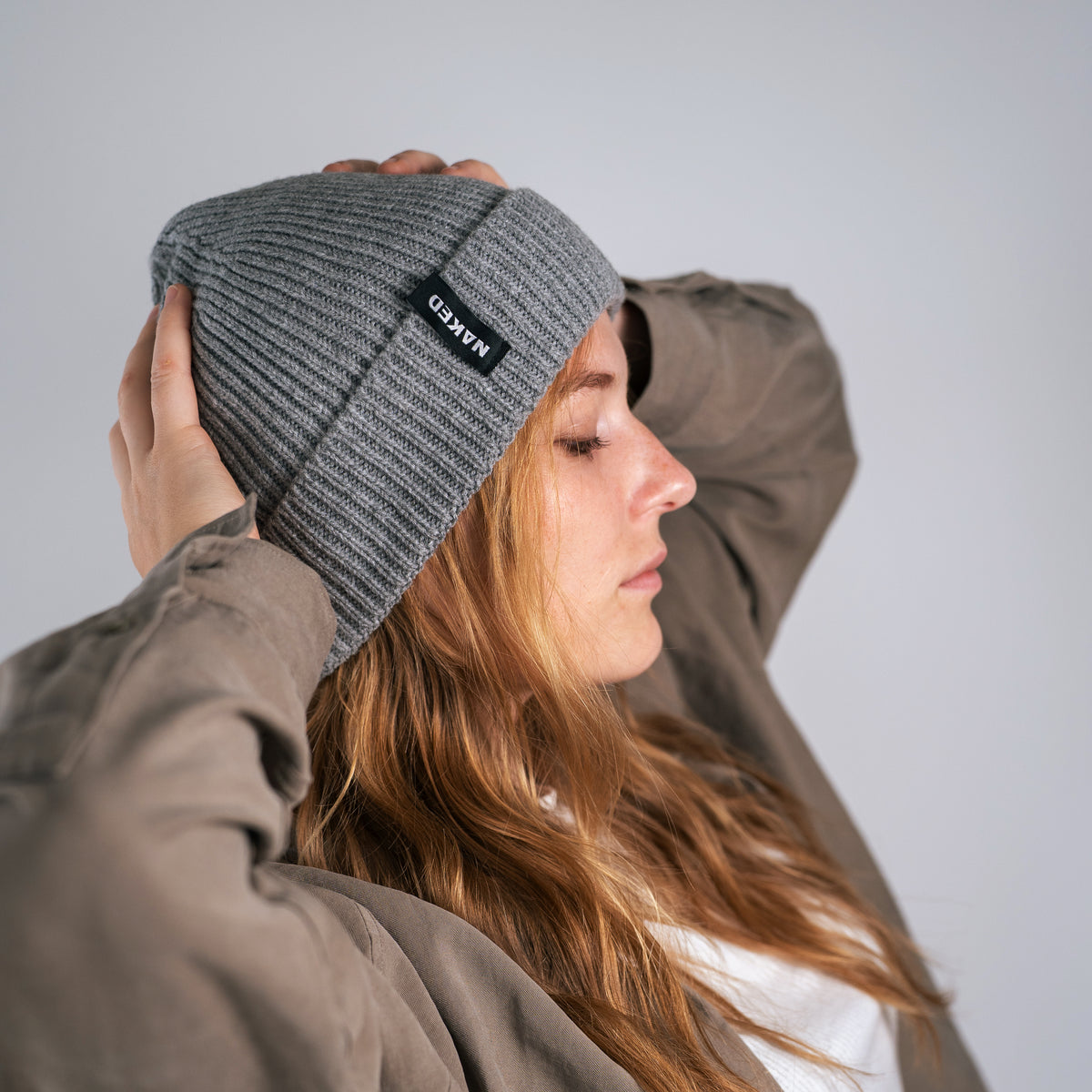 Eine Person mit einer grauen NAKED Optics Beanie POLAR und einer braunen Jacke posiert im Profil und rückt die Mütze vor einem schlichten Hintergrund zurecht.