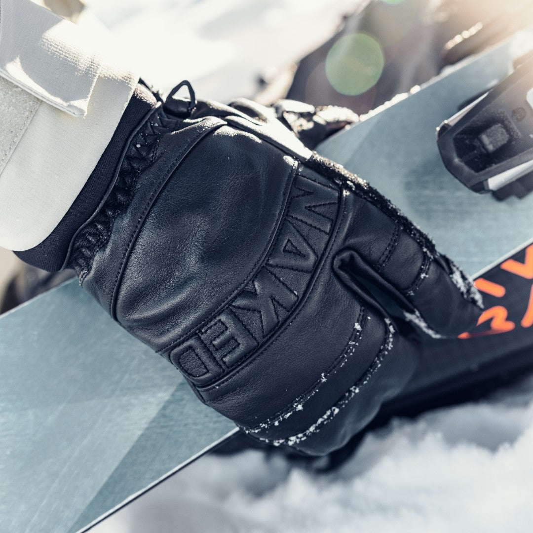 A gloved hand holds a ski, with snow visible on the NAKED Optics CRUZER EVO Mitten and the ground.