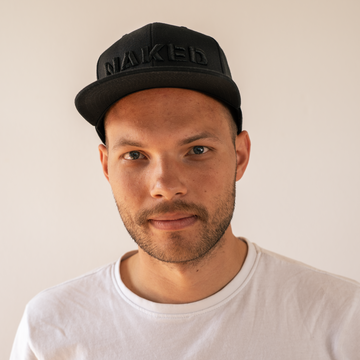 Ein Mann in einem weißen Hemd und einer schwarzen „NAKED Optics“-Snapback-Kappe blickt vor einem schlichten, hellen Hintergrund in die Kamera.
