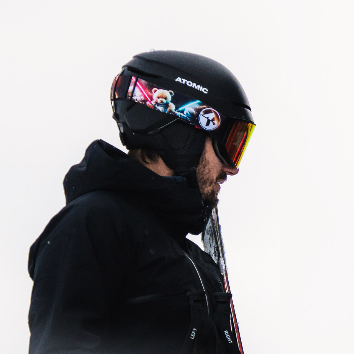 Eine Person in Skikleidung mit einer großen Skibrille, dem Modell „The NOVA“ von NAKED Optics, und einem Bärenaufkleber auf dem Helm steht vor einem weißen Hintergrund und blickt nach rechts. Die Skibrille ist mit einer praktischen Anti-Beschlag-Beschichtung ausgestattet.
