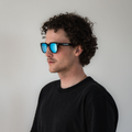 Ein Mann mit lockigem Haar, der eine blaue „The SWOOP“-Sonnenbrille von NAKED Optics mit UV-Schutz und ein schwarzes Hemd trägt und vor einem schlichten weißen Hintergrund steht.