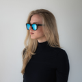 Eine Frau mit langen blonden Haaren trägt die SWOOP-Sonnenbrille mit polarisierten Gläsern von NAKED Optics und einen schwarzen Rollkragenpullover vor einem schlichten weißen Hintergrund. Die stilvolle Sonnenbrille bietet optimalen UV-Schutz und verleiht ihrem klassischen Look gleichzeitig eine moderne Note.