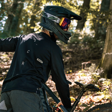 Eine Person, die einen Helm und dunkle Schutzkleidung trägt und die MTB-Brille mit Wechselgläsern von NAKED Optics trägt, steht neben dem TROOP Bike in einem sonnendurchfluteten Waldgebiet.