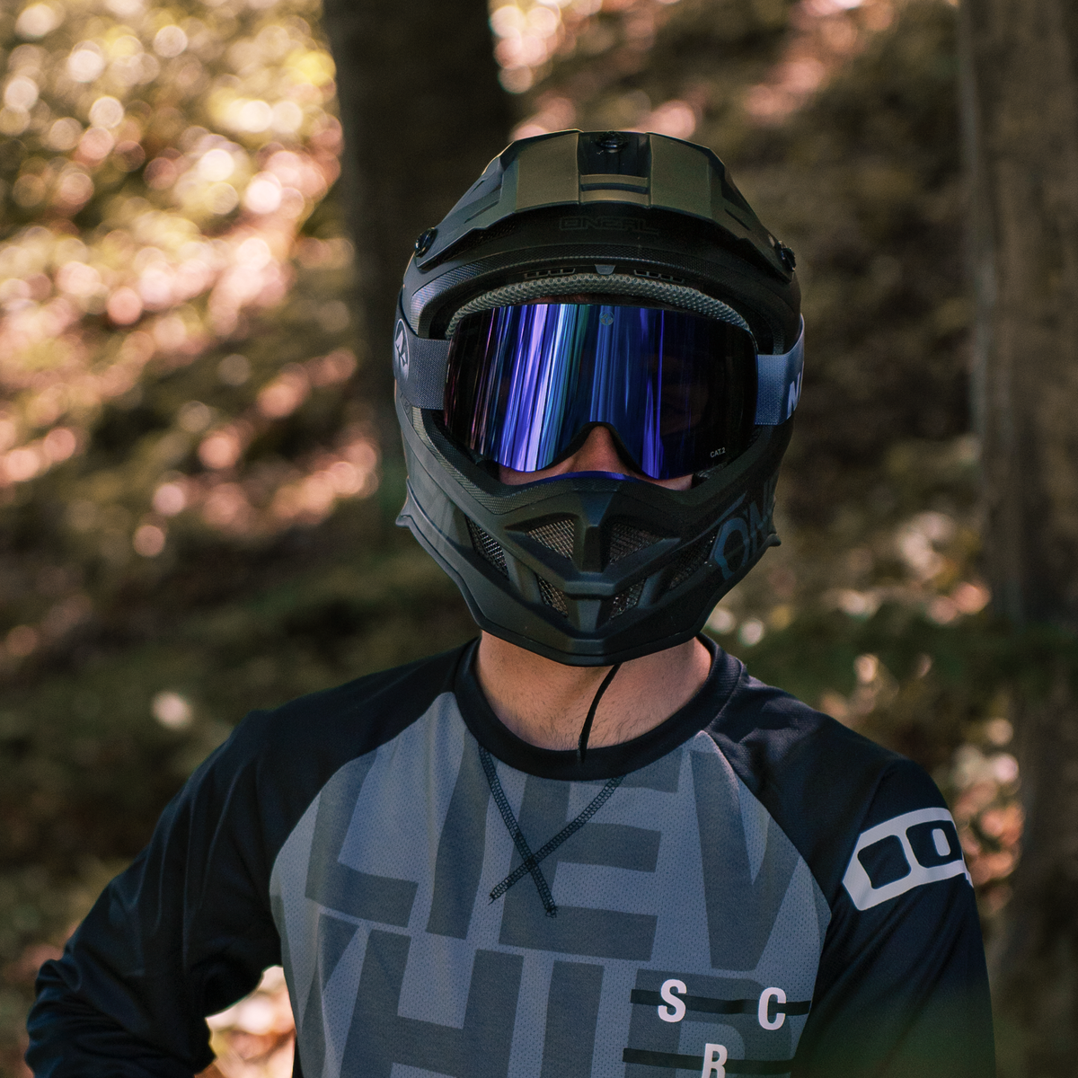 Der Fahrer, gekleidet in ein gemustertes Trikot und mit einem Integralhelm, steht in einem sonnenbeschienenen Wald und trägt „The TROOP Bike“ von NAKED Optics – eine MTB-Brille mit Wechselgläsern für optimale Sicht auf dem Trail.