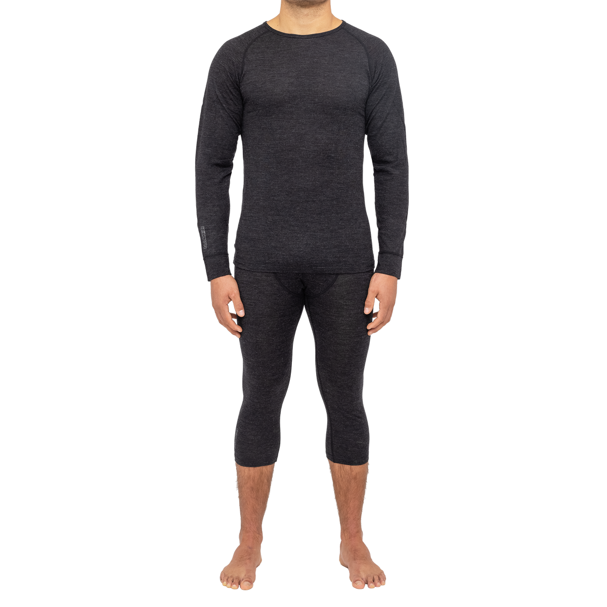 Ein Mann steht barfuß vor einem weißen Hintergrund und trägt ein farblich passendes dunkles Langarm-Thermoshirt sowie eine passende Thermohose. Das Set „NAKED Optics Thermo-Unterwäsche Herren (100 % Merinowolle)“ bietet außergewöhnlichen Tragekomfort und Wärme und eignet sich daher perfekt für den Alltag oder als leistungsstarke Funktionswäsche aus Merinowolle für Outdoor-Abenteuer.