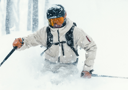 Skifahrer in weißer Jacke und orangefarbener Skibrille fährt durch tiefen Schnee, während es schneit.