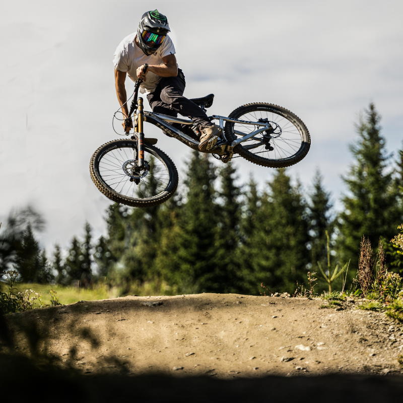 Ein Mountainbiker springt hoch von einem Feldweg ab, im Hintergrund sind Bäume und ein blauer Himmel zu sehen.