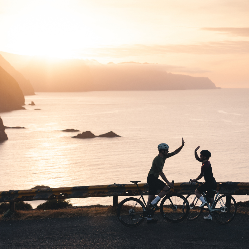 Zwei Radfahrer, die sich bei Sonnenuntergang am Meer ein High Five geben, im Hintergrund sind Klippen und das Wasser zu sehen.
