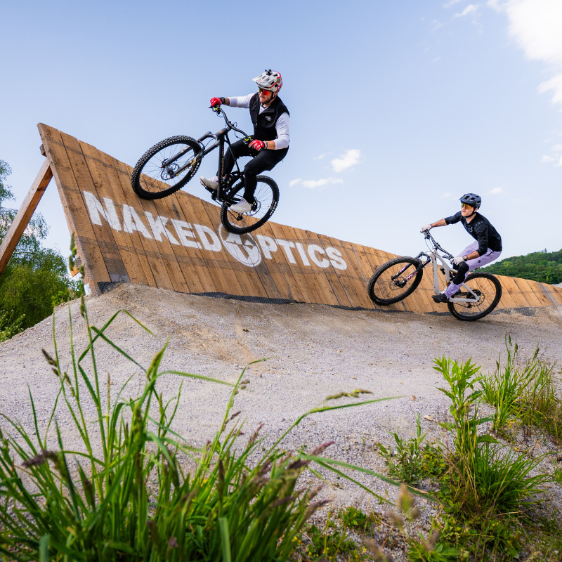 Zwei Radfahrer fahren unter blauem Himmel mit Mountainbikes auf einer schrägen Holzrampe, die mit „NAKED OPTICS“ beschriftet ist.