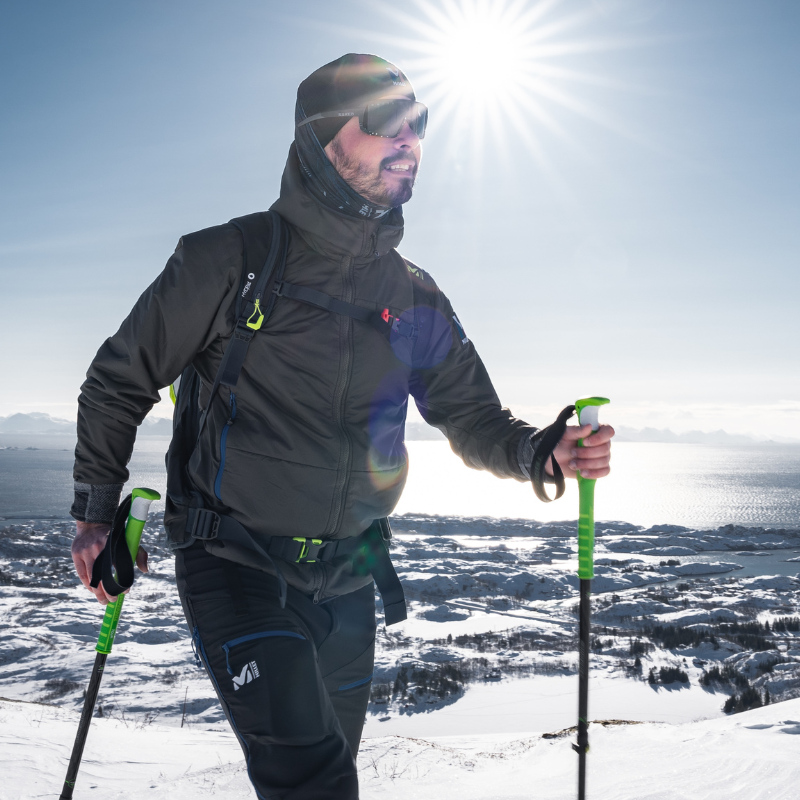 Ein Mann in Winterkleidung, der mit Wanderstöcken auf einem verschneiten Berg wandert, während die Sonne hell scheint.
