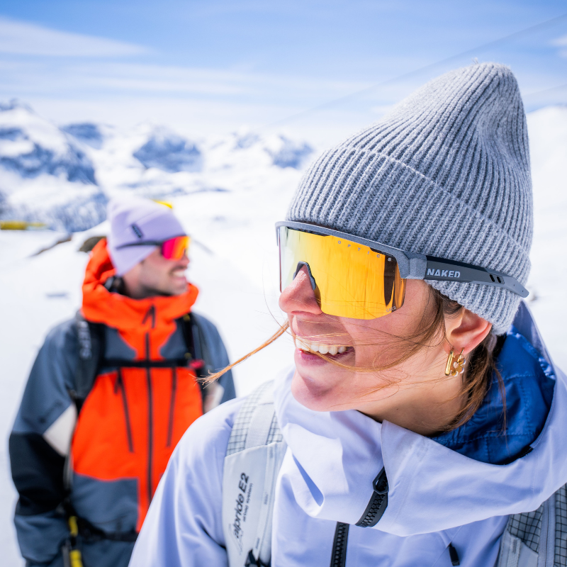Zwei Personen in Skikleidung, die auf einem verschneiten Berg lächeln, mit hellen Sonnenbrillen und Winterkleidung.
