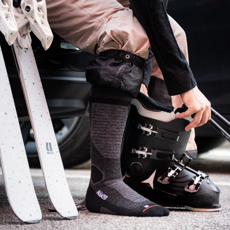 Eine Person, die neben weißen Skiern einen schwarzen Skischuh anzieht und graue Socken sowie eine beige Skihose trägt.