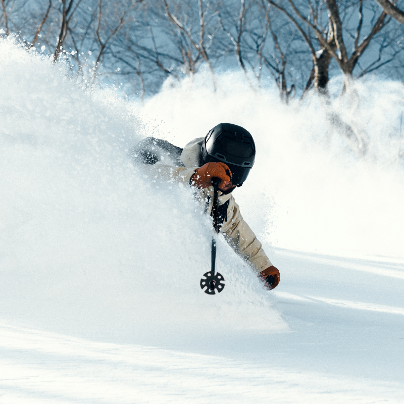 Ein Skifahrer mit Helm, der durch den Tiefschnee carvt, umgeben von einer Pulverschneewolke, im Hintergrund Bäume.