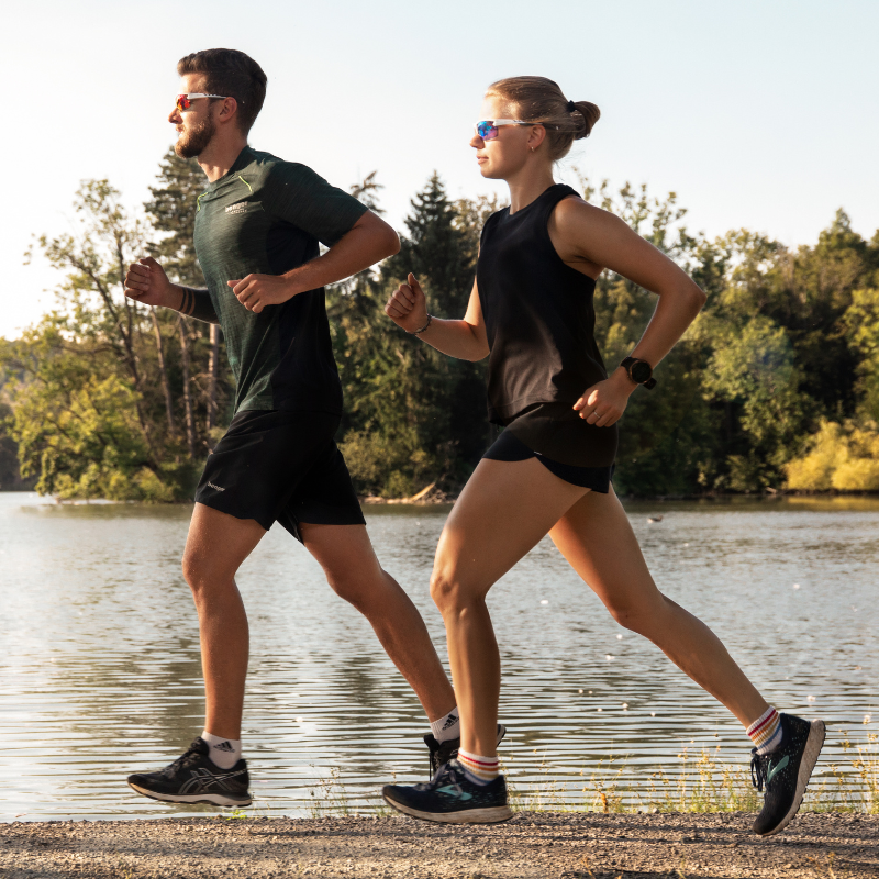 An einem sonnigen Tag joggen zwei Personen in Sportkleidung und mit Sonnenbrillen am See entlang. Im Hintergrund sind Bäume zu sehen.