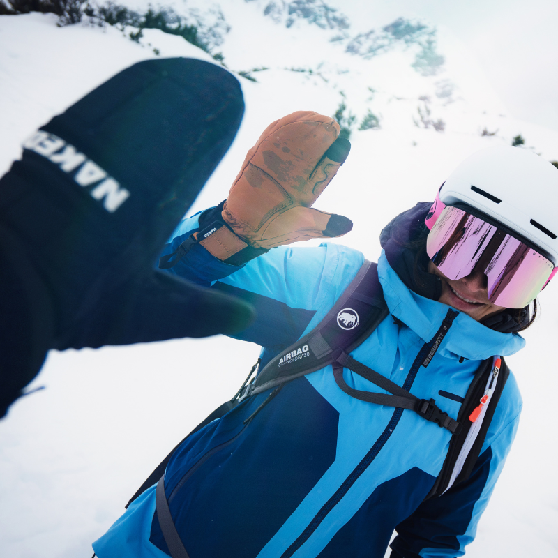 Eine Person in Skikleidung und mit Helm gibt auf einem verschneiten Berghang ein High Five, lächelt und trägt eine Skibrille.