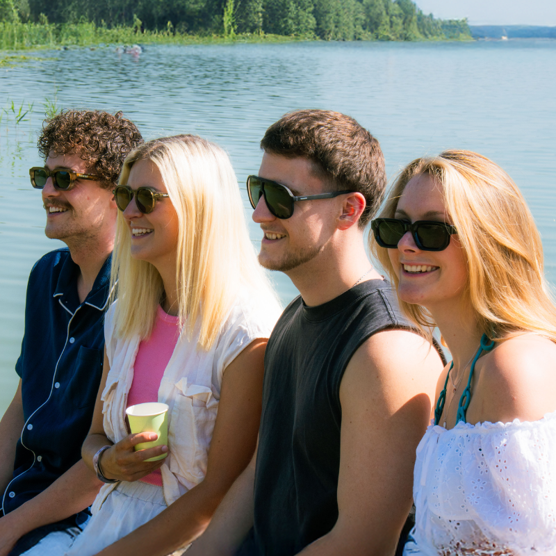 Vier lächelnde Erwachsene mit Sonnenbrillen sitzen an einem sonnigen Tag am See, umgeben von viel Grün.