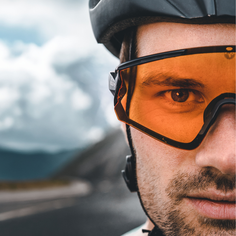 Nahaufnahme des Gesichts eines Radfahrers, der eine orangefarbene Sonnenbrille und einen Helm trägt, im Hintergrund sind die Straße und der Himmel unscharf zu sehen.