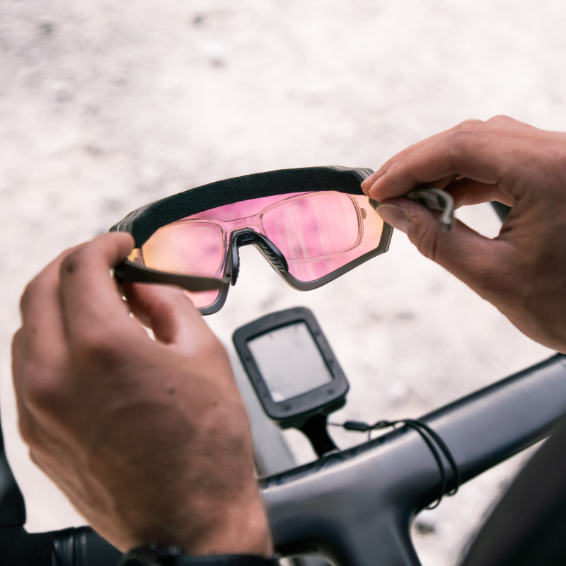 Hände, die eine Fahrradbrille mit rosa Gläsern über einem Fahrradlenker reinigen, an dem ein Fahrradcomputer befestigt ist.