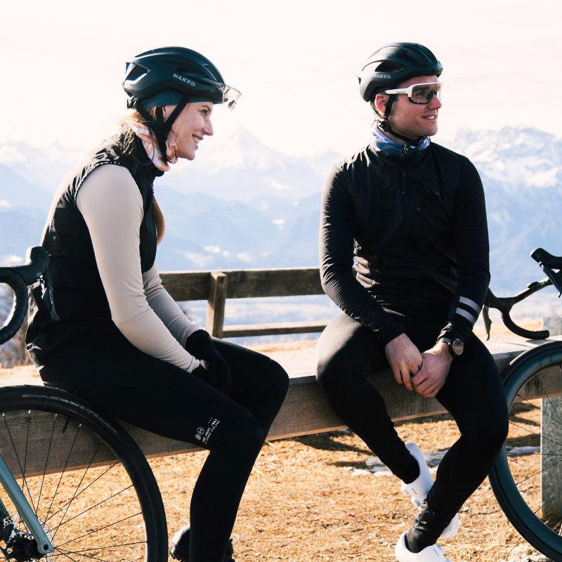 Zwei Radfahrer mit Helmen und Sonnenbrillen sitzen draußen auf einer Bank, im Hintergrund sind Berge zu sehen.