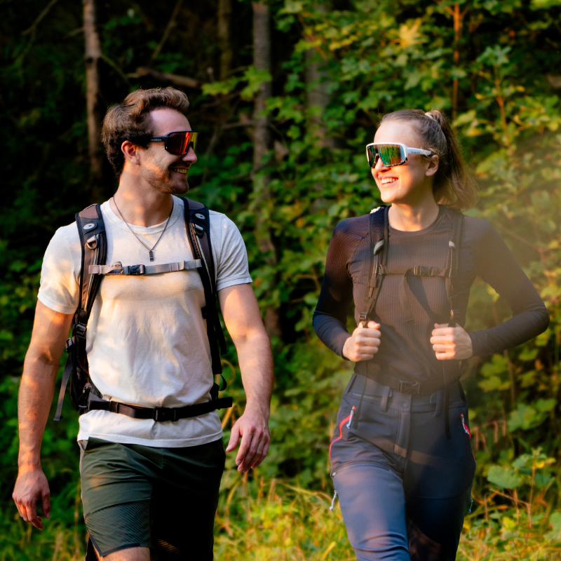 Zwei Menschen mit Sonnenbrillen und Rucksäcken wandern in der Natur und lächeln sich an einem sonnigen Tag vor dem Hintergrund von grünem Laub an.