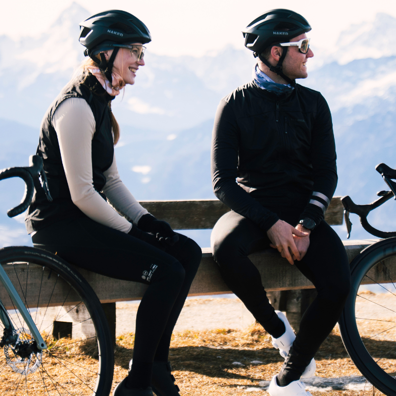 Zwei Radfahrer mit Helmen und Sonnenbrillen sitzen draußen auf einer Bank, im Hintergrund sind Berge zu sehen.