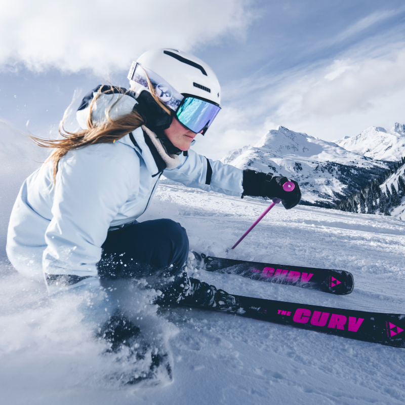Ein Skifahrer in blauer Ausrüstung saust die verschneiten Hänge hinunter, im Hintergrund sind Berge und der Himmel zu sehen.