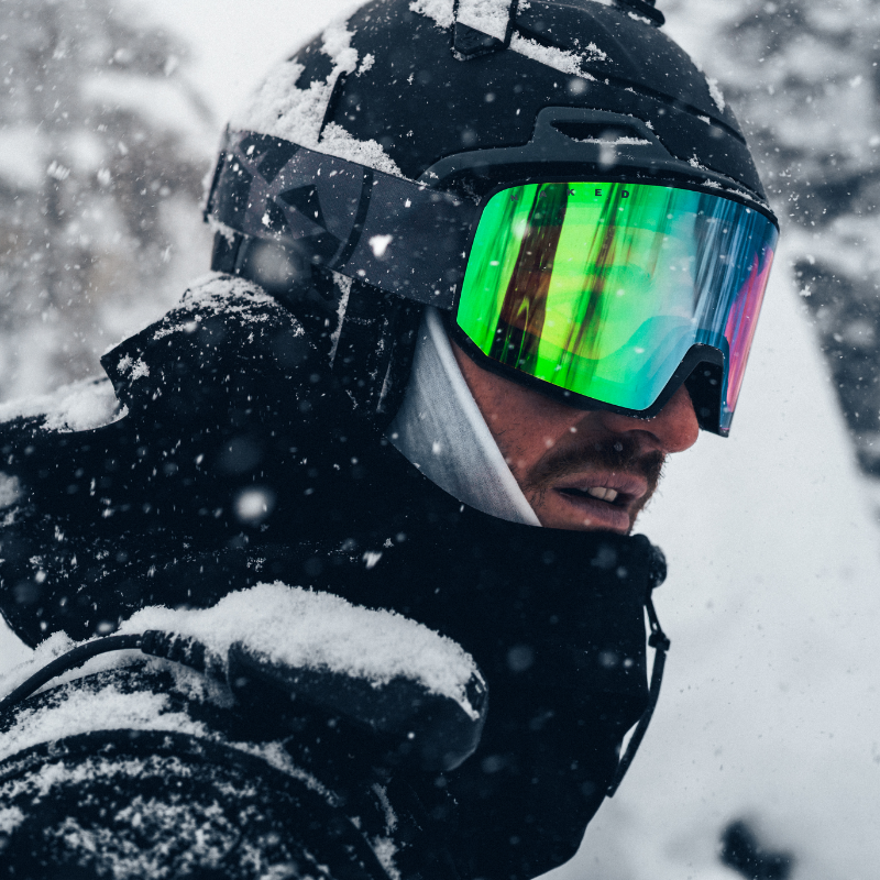 Eine Person in Skikleidung und mit einer reflektierenden Skibrille steht im fallenden Schnee, wobei Helm und Jacke mit Schnee bedeckt sind.