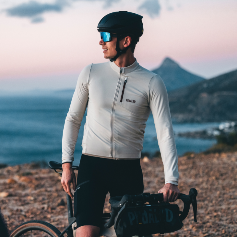 Ein Radfahrer mit Sonnenbrille und Helm steht mit seinem Fahrrad auf einem felsigen Weg, im Hintergrund sind bei Sonnenuntergang das Meer und die Berge zu sehen.
