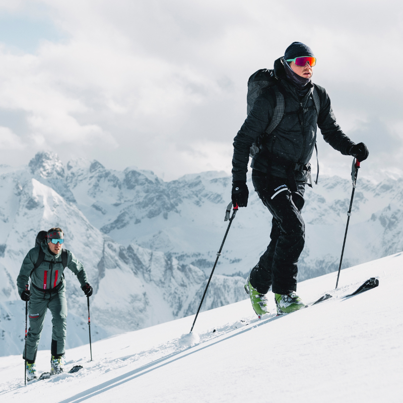 Zwei Personen, die in Winterkleidung und mit Sonnenbrillen auf verschneiten Berghängen bergauf Ski fahren. Im Hintergrund sind schneebedeckte Gipfel zu sehen.