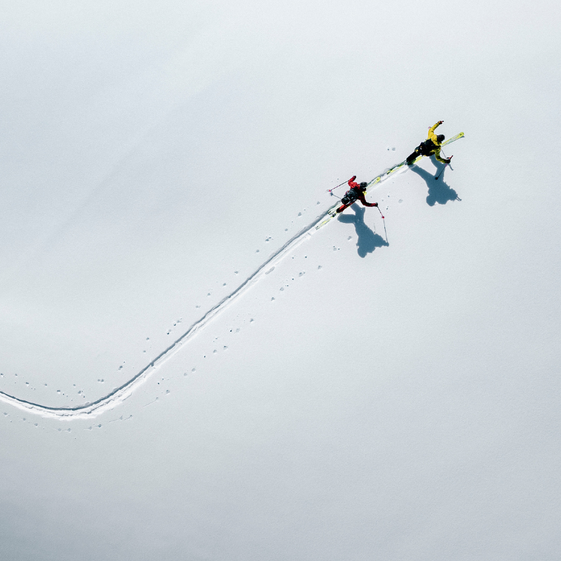 Zwei Skifahrer in bunten Outfits ziehen ihre Spuren durch eine weite, unberührte Schneelandschaft – aus der Vogelperspektive betrachtet.
