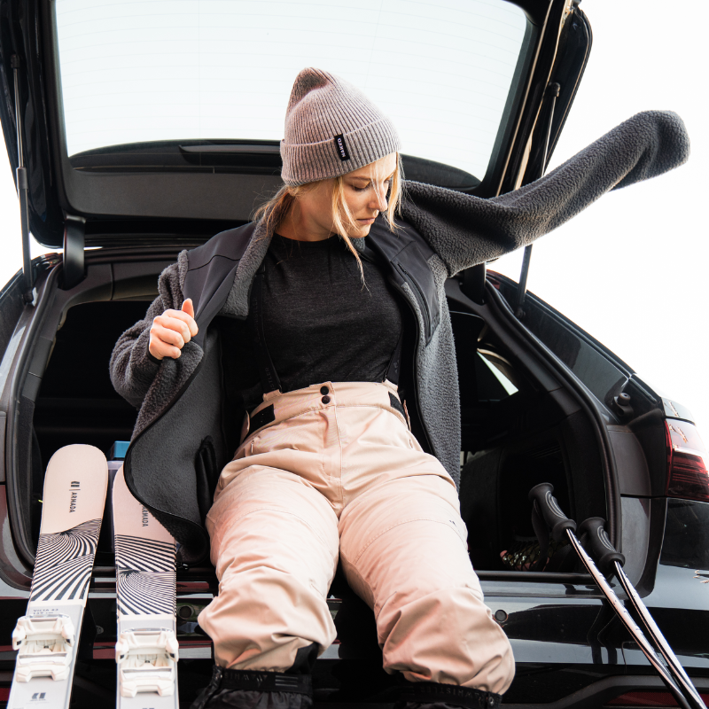Eine Frau sitzt im offenen Kofferraum eines Autos und zieht sich eine Jacke an; neben ihr liegen Skier und Skistöcke.