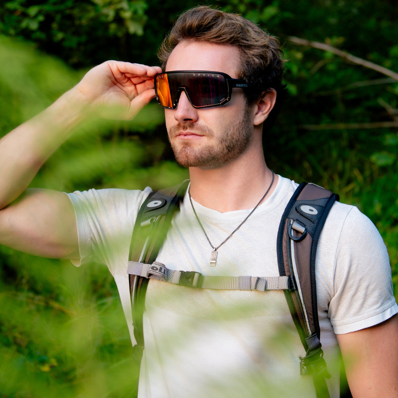 Ein Mann in einem weißen Hemd und mit einem Rucksack rückt sich in einer grünen Außenkulisse eine große Sonnenbrille zurecht.