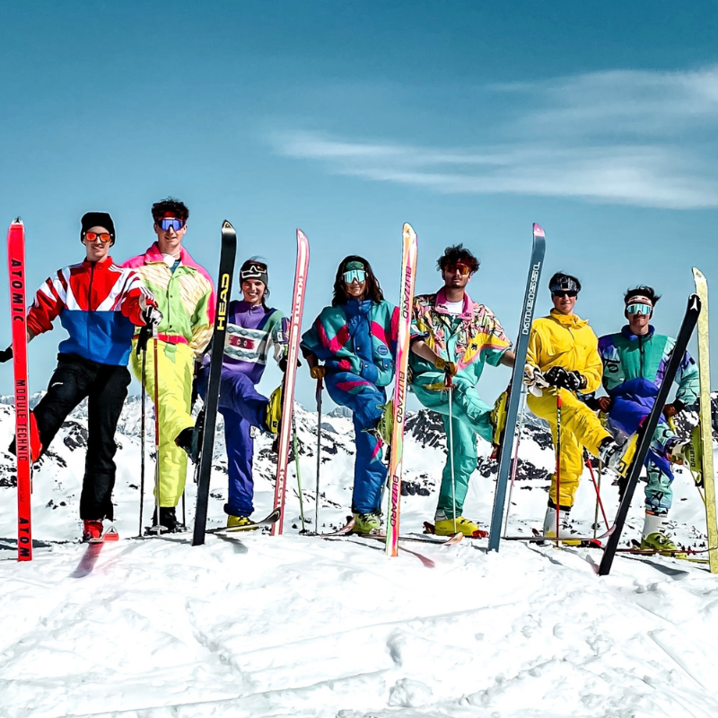 Sieben Menschen in bunten Retro-Skianzügen posieren mit Skiern auf einem verschneiten Berg unter strahlend blauem Himmel.