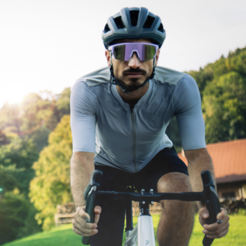 Ein Radfahrer mit Helm und Sonnenbrille fährt im Freien, im Hintergrund sind Bäume und ein Haus zu sehen.