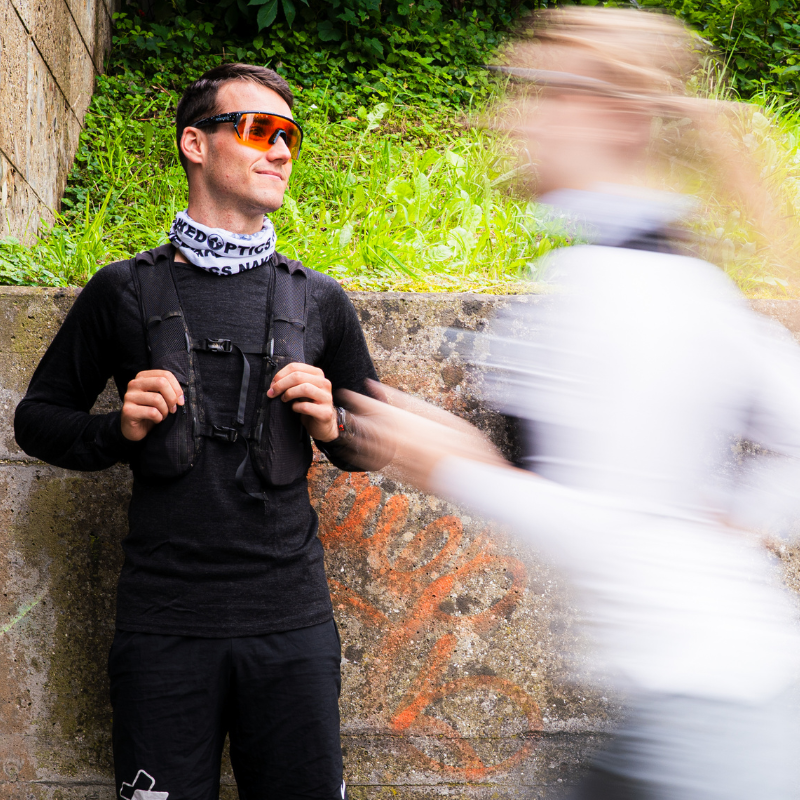 Ein Mann mit Sonnenbrille steht an einer Mauer, während vor ihm im Freien eine verschwommene Gestalt vorbeijoggt.