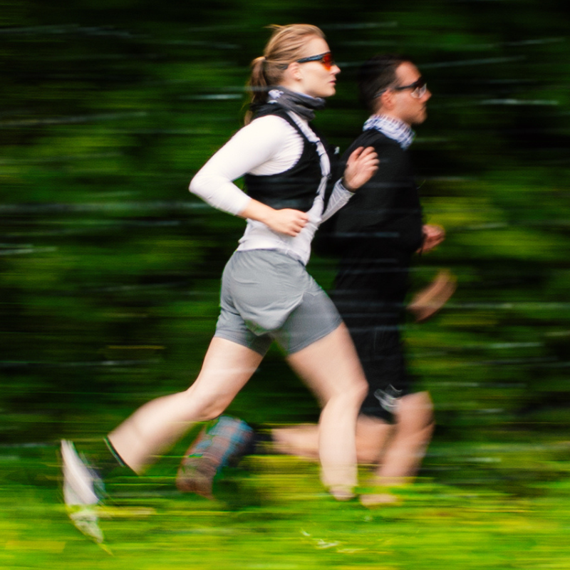 Zwei Personen in Sportkleidung laufen im Freien nebeneinander; Bewegungsunschärfe verdeutlicht ihre Geschwindigkeit, grüner Hintergrund.