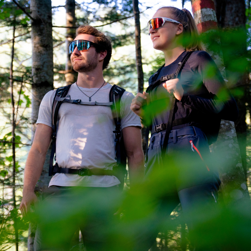 Zwei Menschen mit Sonnenbrillen und Rucksäcken stehen in einem sonnendurchfluteten Wald, umgeben von grünem Laub.
