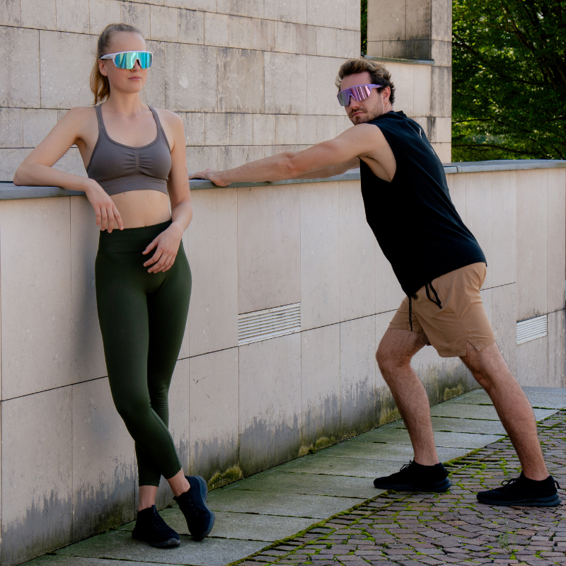 Zwei Personen in Sportkleidung und mit verspiegelten Sonnenbrillen, die sich draußen an einer Steinmauer dehnen.