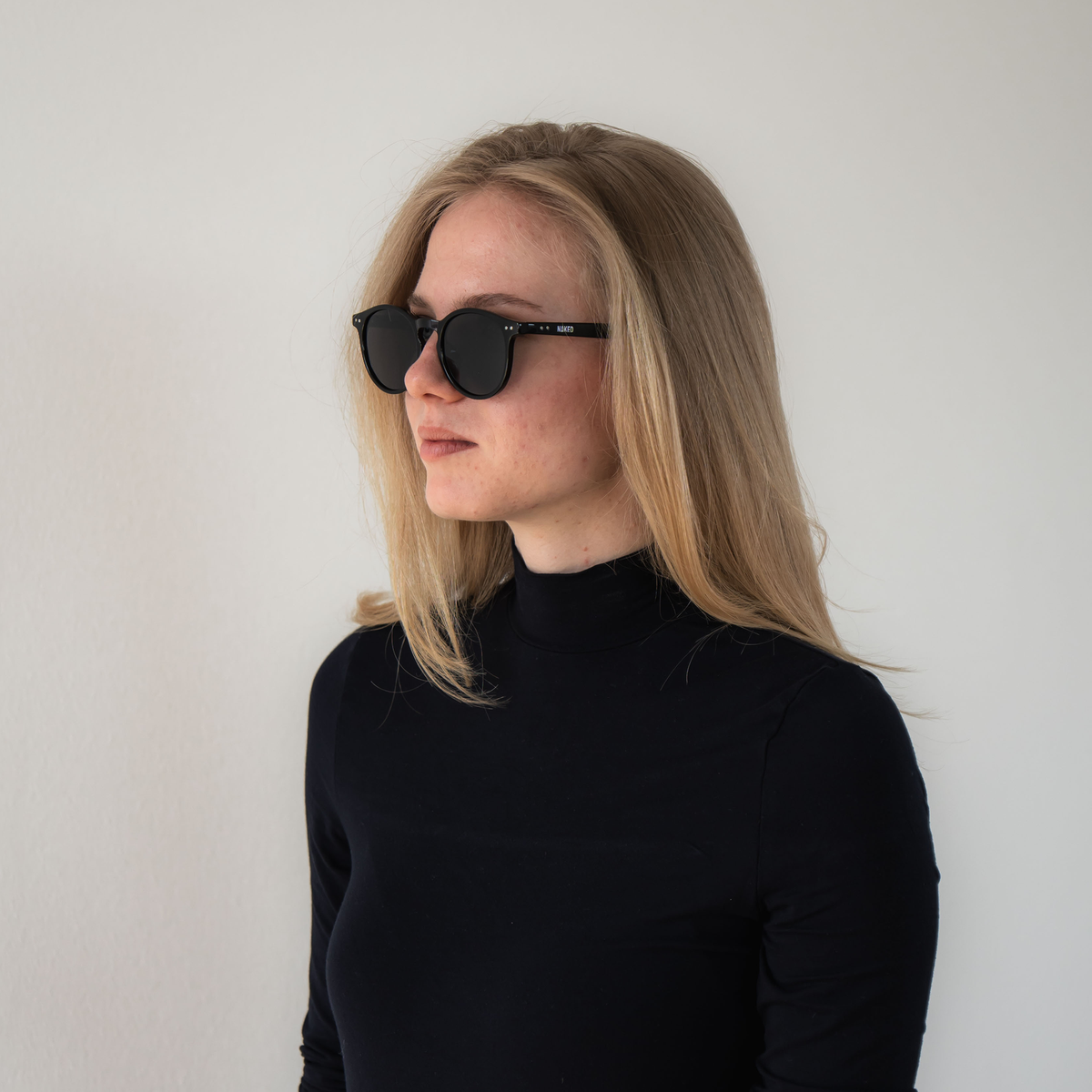 A woman with long blonde hair, wearing a black turtleneck and facing left against a plain wall, sports The WHOOP by NAKED Optics—polarised sunglasses with UV400 protection that offer both stylish looks and optimal eye safety.