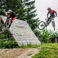 Two mountain bikers in gear, each wearing The RIFT high-performance Downhill Brille with Anti-Fog Glas by NAKED Optics, jump off a wooden ramp surrounded by grass and trees.