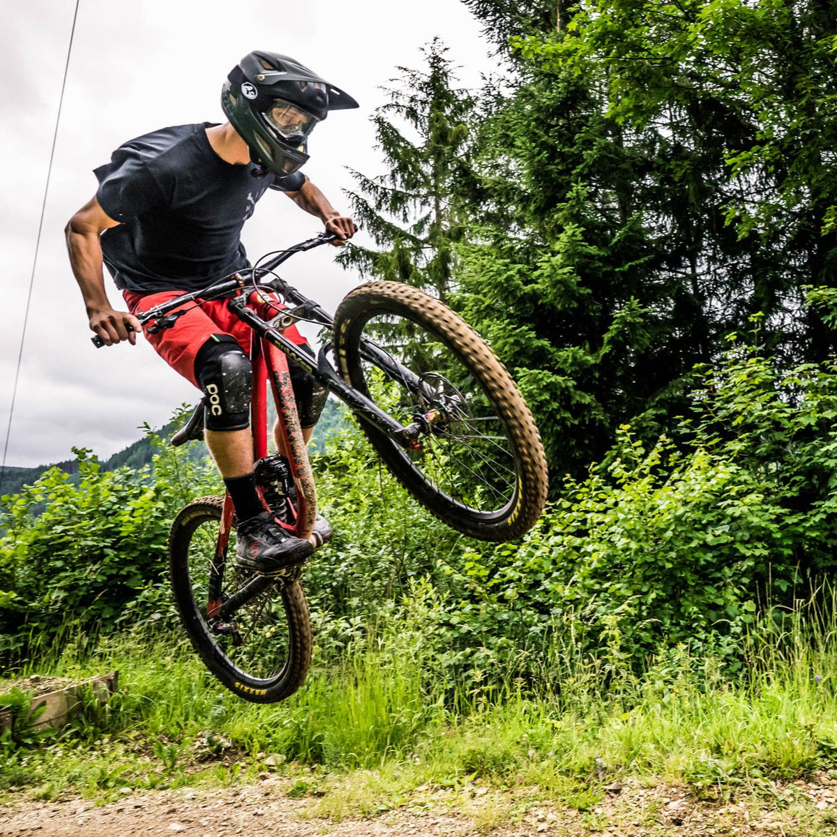 A mountain biker in red shorts launches off a dirt trail amidst lush green trees and foliage, confidently wearing NAKED Optics The RIFT goggles for crystal-clear vision on the ride.