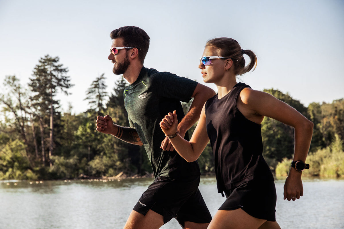 In una giornata di sole, due persone fanno jogging in riva al lago; entrambe indossano gli eleganti occhiali da sole sportivi NAKED Optics The RUSH Halfframe con protezione UV400, che completano i loro outfit sportivi. Sullo sfondo si vedono degli alberi.