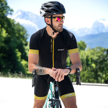 Ein Mann in schwarzer Fahrradbekleidung und mit Helm lehnt sich im Freien an ein Fahrrad. Er trägt die „The RUSH Halfframe“ von NAKED Optics – eine Sportsonnenbrille mit UV400-Schutz – vor dem Hintergrund von Bergen und Bäumen.
